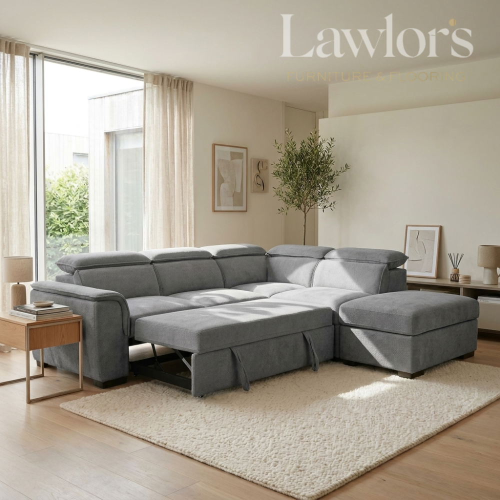 A large gray modular sectional sofa with a chaise lounge and matching ottoman in a bright, modern living room. The sofa is styled on a cream shag rug with a wooden side table next to it.